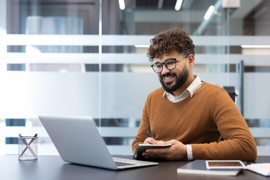 Young man smiling and taking notes in a notebook while working on a laptop computer at a modern office desk, focusing on productivity and professionalism