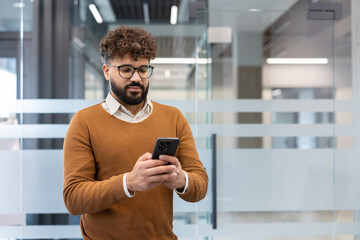 Young businessman with curly hair and beard wearing glasses checks smartphone at a contemporary office desk, texting and browsing apps during a busy workday, focused on screen