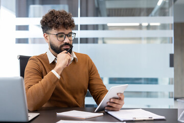 Young man wearing glasses and a beard sitting at a desk, deeply concentrating and thinking while reviewing information on a digital tablet in a modern office environment