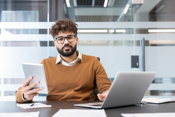 Young professional with glasses and curly hair working on laptop and tablet at a modern office desk, focused on digital tools and productive startup-style business planning