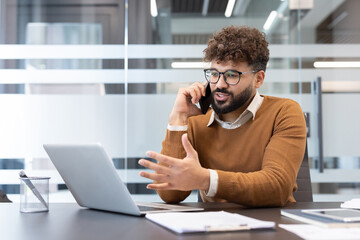 Young man with curly hair and glasses engaged in a phone conversation, working remotely from a contemporary office space with a laptop and documents on his desk