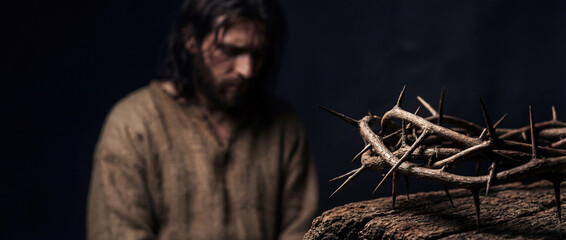 Crown of thorns on wooden surface with blurred Jesus Christ looking down in background. Biblical concept of Passion, suffering and sacrifice. Religious banner for Good Friday and Lent.