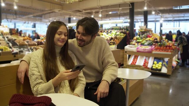 Youthful pair enjoy shopping break, Young lovers rest on market bench, Casual couple pause amid vibrant market stalls, Romantic young pair relax comfortably at busy market with colorful surroundings