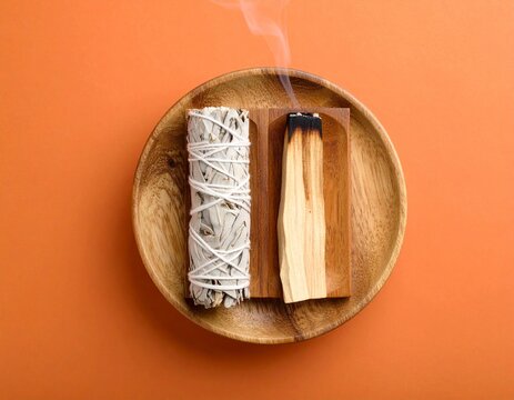 White sage and palo santo sticks are burning in a bundle of incense on a wooden stand against an orange-brown backdrop. Use holy wood sticks for room fumigation and meditation. overview