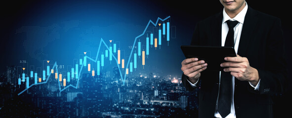 A business professional stands in front of a digital stock market graphic while analyzing data on a tablet, representing finance and urban growth in a modern setting. Gantry