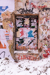 Moscow, Russia, Weathered brick wall with a small window covered in colorful posters and graffiti tags surrounded by peeling plaster and snow at the base. 
