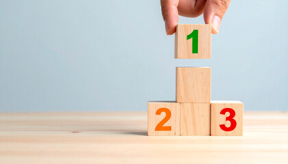 Hand placing a wooden block labeled &ldquo;1&rdquo; atop two stacked blocks labeled &ldquo;2&rdquo; and &ldquo;3&rdquo; featuring a podium. The image symbolizes prioritization, competitiveness, achieving budgets in the business world.