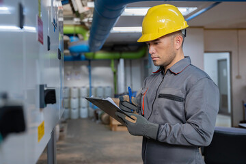 Professional technician inspecting electrical control panel in a factory plant. Male engineer recording operational data on clipboard during safety maintenance of industrial machinery system.