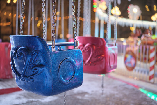 Close up of an empty swing ride seat hanging on chains at a fairground, with colorful night lights and bokeh creating a festive winter atmosphere, ideal for holiday and leisure concepts.