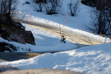Guided passage narrow snow-covered road curving through winter terrain, plowed track marked by tall orange poles, trees and rocks framing shaded light
