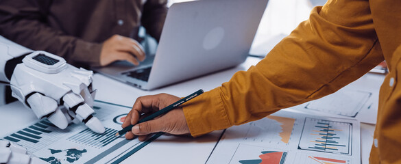 A human hand draws on a chart while a robotic hand rests on the table, representing the synergy of human creativity and advanced technology in a modern office.Noogenesis