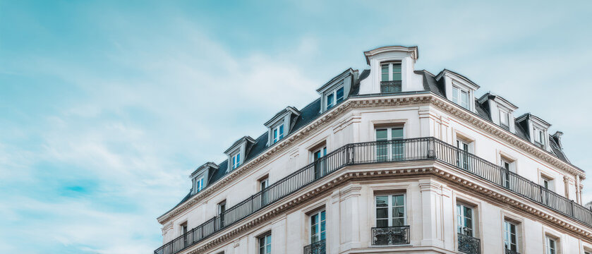 Classic light-colored apartment building with ornate iron balconies and mansard roof, photographed under a bright blue sky.
