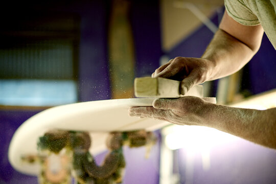 Surfboard, hands and sanding in workshop for manufacturing, design or polish. Surf, person and tools to shape blank with block, smooth and production process with dust on foam for creative handcraft