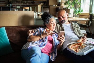 Mature couple laughing over smartphone while eating pizza at home