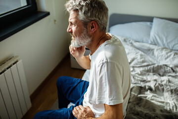 Mature man looking pensive while sitting on bed in bedroom