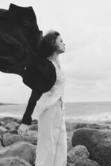 Wind swept, white outfit flares against a rocky shoreline in monochrome, a dramatic sea backdrop...