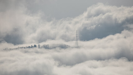 Winter mountain dawn, where a high-voltage pylon rises through rolling clouds and fog, silhouetted against soft morning light.