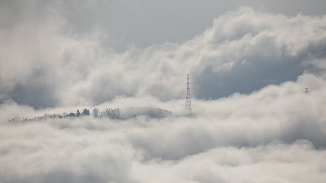 Winter mountain dawn, where a high-voltage pylon rises through rolling clouds and fog, silhouetted against soft morning light. - Powered by Adobe