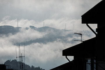 Winter mountain dawn, where a high-voltage pylon rises through rolling clouds and fog, silhouetted against soft morning light.