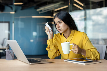 Sick young Indian woman sitting at a desk in the office, holding a napkin and a cup, feeling unwell