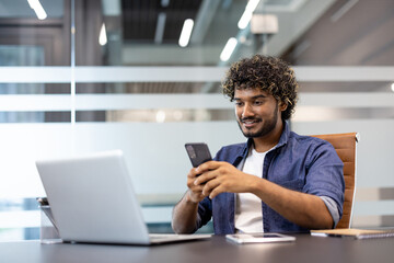 Smiling young Indian man working in an office, sitting at a desk and using a mobile phone