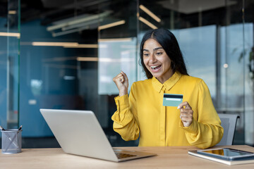 A happy young businesswoman is sitting at a desk in the office, holding a credit card and looking at the laptop screen with satisfaction
