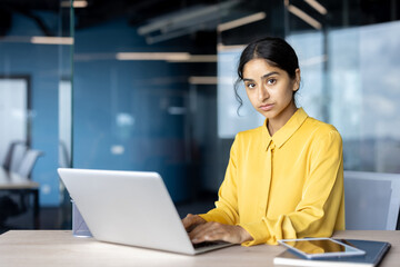 Portrait of a young serious businesswoman in a yellow shirt sitting at a desk in the office, working on a laptop and looking at the camera