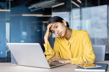 Tired young Indian businesswoman sitting tiredly at office desk holding her head