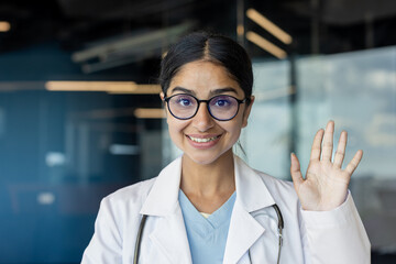Close-up portrait of a young Indian female doctor in a hospital wearing a white coat, standing in front of the camera, greeting and waving