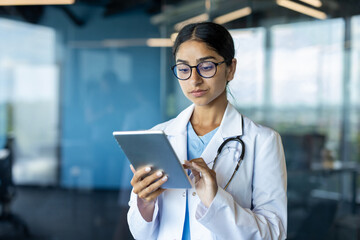 Young Indian female doctor in white coat standing in clinic room and using tablet