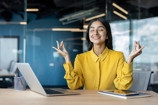 Relaxed young Indian businesswoman sitting at her desk in the office in lotus position, resting and meditating