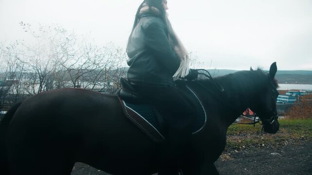 A long shot of an adult woman wearing a jacket and scarf, sitting astride a black horse, holding the reins. They walk against the backdrop of an autumn landscape and an overcast sky in countryside