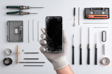 Hand holding a cracked smartphone surrounded by various repair tools on a clean surface, illustrating the process of mobile device repair and maintenance techniques