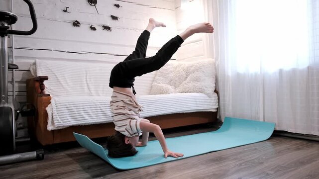 Caucasian boy doing headstand on yoga mat in the home interior. Nine year sporty child. Hip-hop and breakdance dancer. Kid does gymnastic exercises. Yoga practice. Sport practicing. HQ 4k footage.