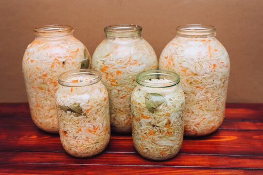 Fresh homemade sauerkraut in glass jars of various sizes on a brown wooden table with a beige background. Concept of fermented foods and natural probiotics.