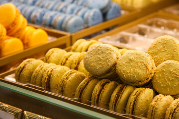 Colorful assortment of French macarons displayed in a bakery showcase. The image features vibrant yellow, blue, pink, and pastel macarons arranged in neat rows, highlighting texture and detail