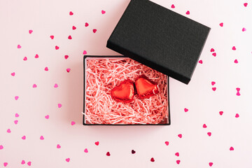 Open black gift box with red heart chocolates inside on pink background with scattered confetti. Valentines day concept. Top view, flat lay