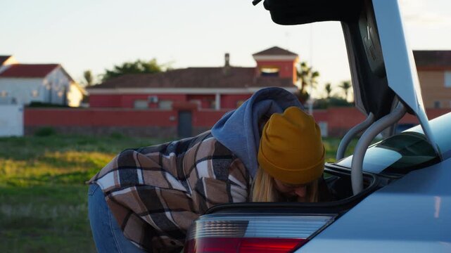Young blonde woman in yellow beanie and plaid shirt searches through an open car trunk at sunset in suburban neighborhood, packing or looking for items during roadtrip