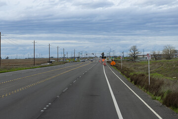 In a rural area, road construction is taking place near a highway. The scene features traffic navigating around the work zone under a cloudy sky during the day