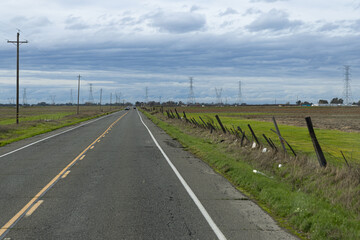 A long, open road extends far into the horizon, running parallel to lush farmland beneath a cloudy sky. In the distance, electric towers rise while a lone car travels along the stretch of asphalt