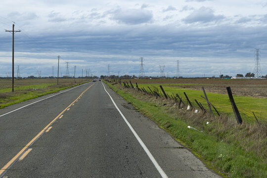 A long, open road extends far into the horizon, running parallel to lush farmland beneath a cloudy sky. In the distance, electric towers rise while a lone car travels along the stretch of asphalt