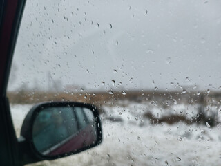 Obraz premium Raindrops collecting on the car window blur the view of the snowy winter landscape. The car's side mirror reflects the scene under the gray sky, creating a gloomy atmosphere.