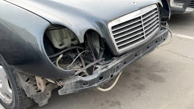 Damaged Car Front End Exposed Bumper Parked On Cracked Asphalt In Urban Lot, Missing Headlights, Twisted Metal, Scratched Paint, Exposed Wiring, Bent Grille, Adjacent Sedan Visible.