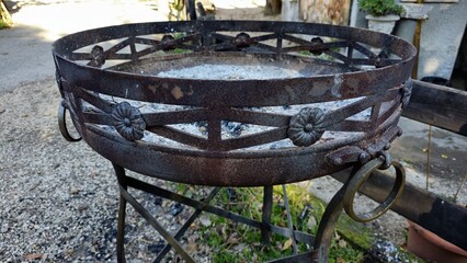 Rome, Italy - 14 January 2025. A rusted iron brazier with flower-shaped accents holds ash, positioned on gravel near a fence and stone wall in a residential outdoor space.