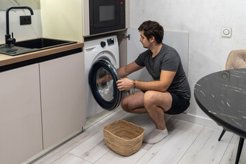 Young man opening front-load washing machine near wicker basket