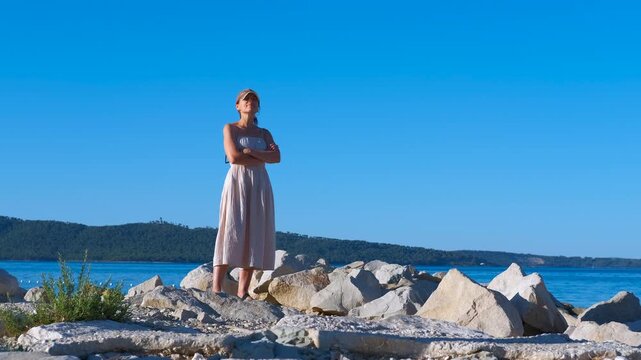 Woman enjoying the summer sun on a rocky beach. Beautiful woman in a long dress and visor cap standing on large rocks by the sea on a sunny day, enjoying the warm weather and beautiful view