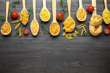 Different types of raw pasta in spoons, tomatoes, rosemary and thyme on wooden table, flat lay. Space for text