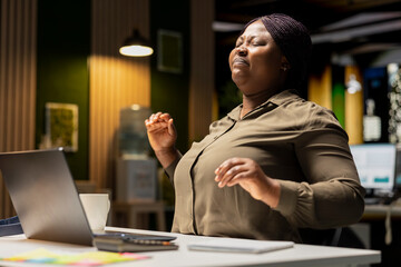 African american woman stretching her upper and lower back muscles, correcting her posture on the chair for muscle relief and relaxation. Feeling discomfort due to intense office activity.