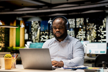 Obraz premium Black person managing daily tasks after hours with focus music on headset, finding inspiration with ambience sounds to help with concentration and productivity. Male worker multitasking.
