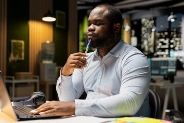 Black young man contemplating about his decisions and project outcomes, searching for possible solutions with laptop after hours. Showcasing intense concentration and logical deduction.